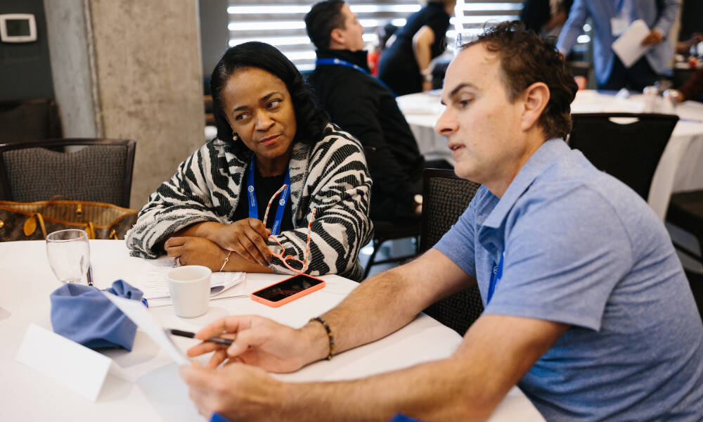 A man and woman talk to each other while sitting at a table during a Board Presidents Leadership Retreat